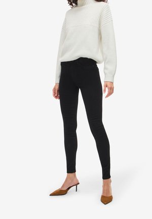 Femme portant un pull à col roulé côtelé blanc, un jean skinny noir, et des mules à talons pointues marron, debout sur fond blanc.