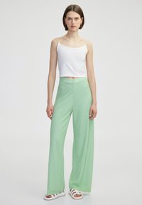 Witte, kortere top met spaghetti bandjes, gecombineerd met lichtgroene, wijde broek met een textuurpatroon. Model draagt witte sandalen.