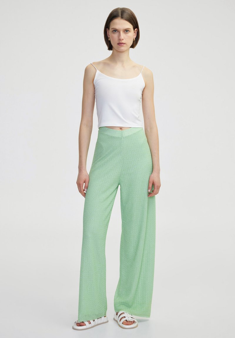 Bílý top na tenké ramínka s krátkým střihem, který je kombinován se světle zelenými širokými kalhotami s texturovaným vzorem. Modelka má na sobě bílé sandály.
