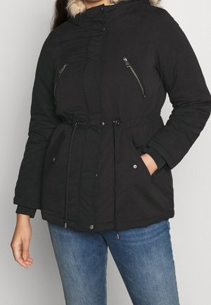 Kobieta w czarnej zimowej kurtce z kapturem obszytym futerkiem, zamkiem błyskawicznym i kieszeniami na zatrzaski oraz ze ściągaczem w talii, ubrana w niebieskie dżinsy.