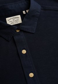 Marineblå skjorte med krave, ribbet tekstur, med gule knapper og et broderet logo samt en vævet vaskeanvisning fastgjort.