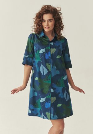 Kvinde med krøllet hår iført en blå og grøn blomsterknapkjole med trekvartærmer, stående mod en ensartet lys baggrund.