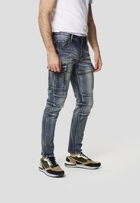 Přiléhavé modré džíny s viditelným prošitím a více kapsami; doplněné o hnědobílé a černé tenisky se sportovním designem.