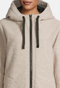 Helle beige Fleecejacke mit strukturierter Oberfläche, ausgestattet mit einem durchgehenden Reißverschluss, verstellbarem Kordelzug an der Kapuze und kontrastierenden dunklen Reißverschlussdetails.