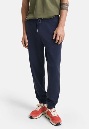 Donkerblauwe sweatpants gemaakt van zachte stof, met een elastische tailleband met trekkoord en taps toelopende boorden. Gecombineerd met rode sneakers.