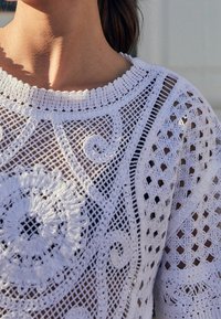 Suéter blanco de ganchillo con patrones intrincados, que incluyen espirales y diamantes, con un cuello de canalé y un acabado texturizado.