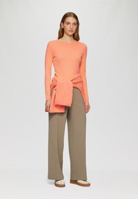 Koraalkleurig longsleeve top met een geknoopte terracotta sjaal om de taille, gecombineerd met wijde beige broeken en tan instap schoenen.