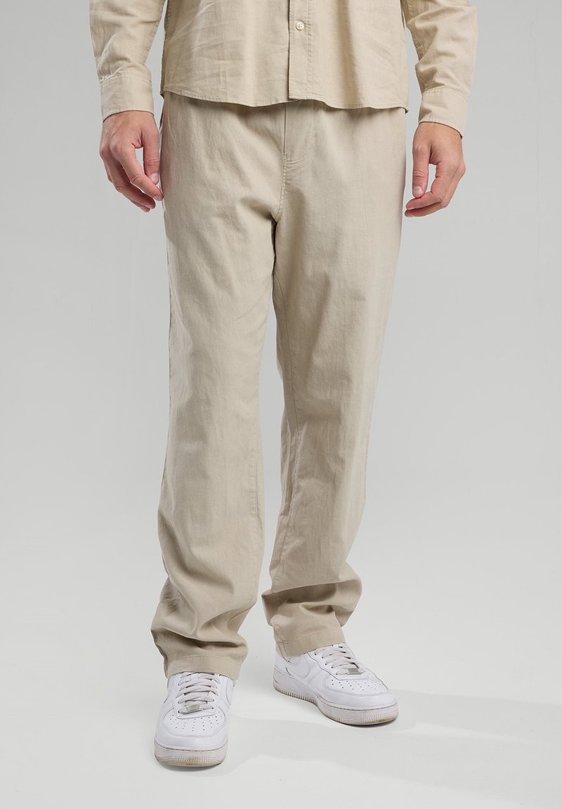 Beige puuvillapantalonit, joissa on rento malli, sivutaskut ja sileä pinta, yhdistettynä valkoisiin tenniskenkiin kontrastiksi.