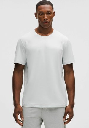 Man with short hair wearing a plain white short-sleeve T-shirt and light gray pants standing against a light background.