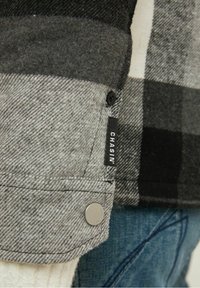 Koszula w kratę w odcieniach szarości i czerni, wykonana z teksturowanej tkaniny, zapinana na srebrne guziki, z metką "CHASIN'" przy mankiecie.