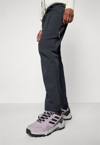 Pantalons de randonnée bleu marine avec poches zippées, coupe ajustée et coutures renforcées. Portés avec des chaussures de sport violet clair avec des accents noirs.