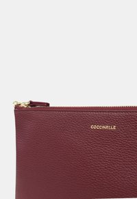 Pochette in pelle texturizzata bordeaux con zip dorata e logo "COCCINELLE" in oro impresso sulla parte anteriore vicino al bordo superiore.