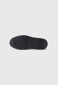 Suela de zapato de goma negra con un patrón de relieve profundo y un logo circular. Superficie texturizada para agarre, que presenta múltiples elementos de tracción elevados.