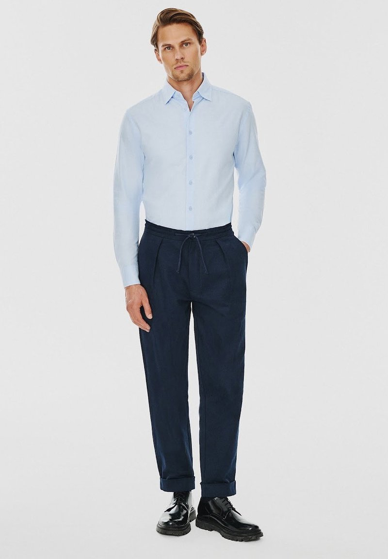 Jasnoniebieska koszula zapinana na guziki, ciemnogranatowe spodnie z elastycznym pasem i czarne buty. Gładki materiał, dopasowany krój, podwinięte mankiety spodni.