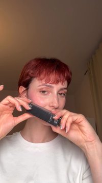 Person with short red hair holding a black KIKO Milano lipstick box near face against a neutral indoor background.