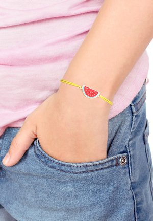 Gelbe Schnurarmband mit einem Wassermelonenanhänger mit roten und grünen Details, getragen am Handgelenk vor rosa Stoff und Denim.