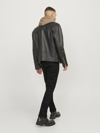 Homme s'éloignant portant une veste en cuir noire, un sweat à capuche marron, un pantalon noir et des bottes noires sur un fond clair uni.