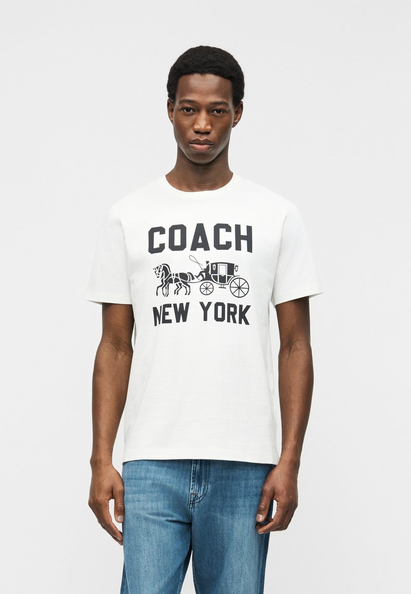 Homme portant un t-shirt blanc avec un graphisme de calèche noire et le texte "COACH NEW YORK", associé à un jean bleu, sur un fond uni.
