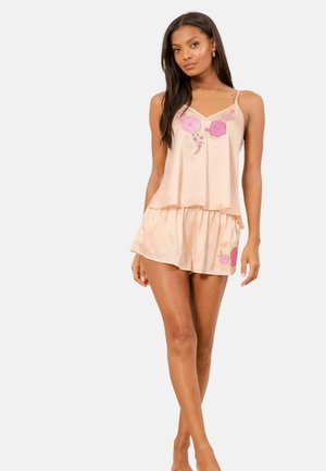 Woman wearing light pink satin camisole and shorts with pink floral embroidery, standing against a plain white background.