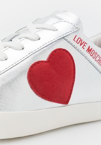 Zilverkleurige leren sneaker met een rode suède hartapplicatie en witte veters; "LOVE MOSCHINO" in rood gedrukt aan de zijkant.