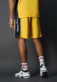 Conjunto deportivo Champion que incluye una camiseta amarilla y pantalones cortos amarillos a juego con detalles en azul marino. Combinado con zapatillas blancas y calcetines a rayas.