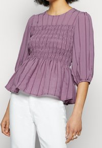 Blouse peplum lavande avec smocks, présentant des rayures verticales et des manches bouffantes. Tissu léger avec une texture douce.