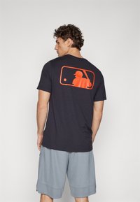 Juodas trumpomis rankovėmis marškinėliai su oranžine beisbolo siluetu atspausdintu ant nugaros. Suderinti su šviesiai pilkais sportiniais šortais.