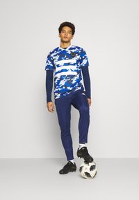 Blauw-wit gestreept atletiekshirt met een camouflagedesign, gecombineerd met marineblauwe broek. De speler staat op een voetbalschoen en draagt zwarte voetbalschoenen.