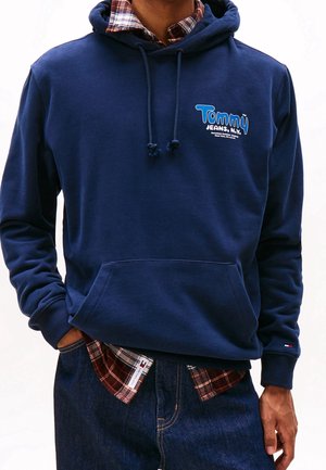Man draagt een marineblauwe Tommy Jeans hoodie over een rood geruit overhemd en donkerblauwe jeans, met handen in de zakken, tegen een effen achtergrond.