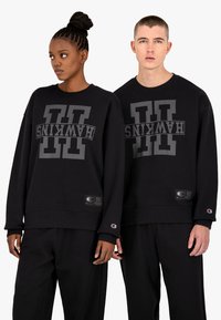 Sudaderas negras con un gran logotipo gris de "HAWKINS". Presenta un cuello redondo acanalado y puños, con parches de la marca cerca del dobladillo.