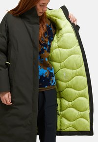 Person, der das hellgrüne, gesteppten Innenfutter eines langen schwarzen Mantels zeigt, trägt einen fleece Pullover in blau-braunem Muster und schwarze Hosen.