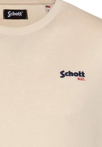 Camiseta de algodón beige con cuello redondo; presenta un logo bordado en azul oscuro de "Schott" y un acento en rojo de "N.Y.C." en el lado izquierdo del pecho.