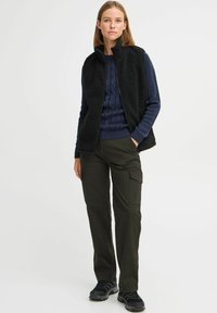 Zwart fleece vest over een marineblauwe kabelgebreide sweater, gecombineerd met olijfgroene cargobroek en zwarte sneakers. Eenvoudig, functioneel ontwerp met praktische details.