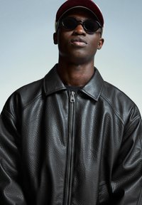 Giacca di pelle nera con finitura texturizzata, dotata di colletto alto e zip frontale. Indossata con occhiali da sole e un cappellino bordeaux.