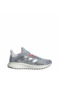 Chaussure de sport grise basse avec une semelle intermédiaire blanche, une semelle extérieure noire, des bandes argentées sur le côté et un détail œillet rouge près des lacets.