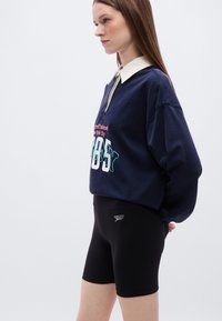 Μπλε ναυτικό φούτερ με λευκό γιακά, άνετη μεγάλη εφαρμογή και τυπωμένο κείμενο· συνδυασμένο με μαύρα, εφαρμοστά σορτς που διαθέτουν λογότυπο κοντά στη μέση.