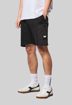 Uomo con pantaloncini neri con tasca laterale a motivo a griglia, sneakers bianche e calzini bianchi con testo nero su sfondo grigio.