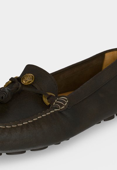 Mocassin en cuir noir avec coutures blanches, nœud décoratif et détail de médaillon doré sur un fond clair.