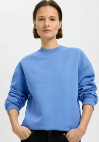 Světle modrý sweatshirt vyrobený z měkké látky, s kulatým výstřihem, volným střihem a žebrovanými manžetami, kombinovaný s černými džíny.