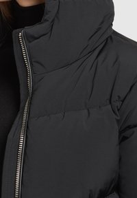 Doudoune noire avec un col haut, dotée d'une fermeture éclair argentée et de détails cousus. Le matériau semble lisse avec une texture douce.