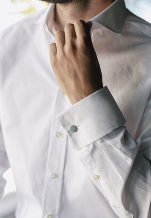 Chemise blanche à col pointu, ornée d'un bouton de manchette gris sur le poignet. Le tissu présente une texture lisse et est impeccablement boutonné.