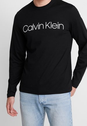 Homme portant un t-shirt noir à manches longues avec le texte blanc « Calvin Klein » et un jean bleu clair sur un fond uni.
