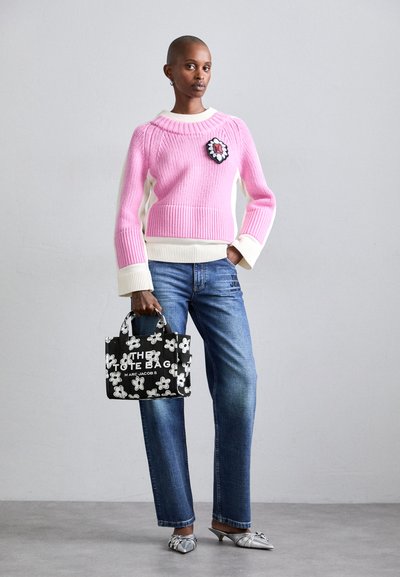 Pull en tricot rose et blanc avec une broche florale, jean droit bleu clair et sac fourre-tout noir avec imprimé floral blanc et texte. Chaussures argentées.