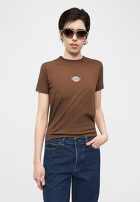 Γυναίκα με κοντά σκούρα μαλλιά που φοράει καφέ μπλουζάκι Dickies, σκουρόχρωμα γυαλιά ηλίου, μπλε τζιν με το ένα χέρι στην τσέπη, σε απλό λευκό φόντο.