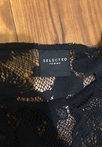 Svart spets tyg med ett blommönster och en etikett som säger "SELECTED FEMME." Texturen är öppen och delikat.