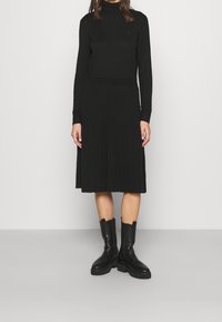 Robe en tricot noire avec un col montant, texture côtelée et manches longues, associée à des bottes cheville noires à semelle épaisse.