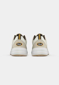 Zapatillas beige con suelas blancas, que presentan detalles marrones, una franja amarilla en la parte posterior y un destacado logo de FILA en el costado.