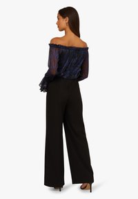 Kobieta z długimi ciemnymi włosami, ubrana w przezroczystą granatową bluzkę na ramiączkach i czarne szerokie spodnie z wysokim stanem, stoi w profilu na białym tle.