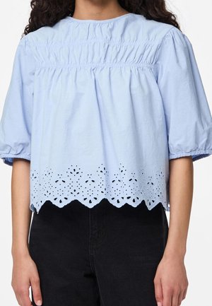 Vrouw draagt een lichtblauw, kort mouwshirt met een geplooide inzet en een bloemenmotief met uitgestanste zoom, gecombineerd met zwarte broek.