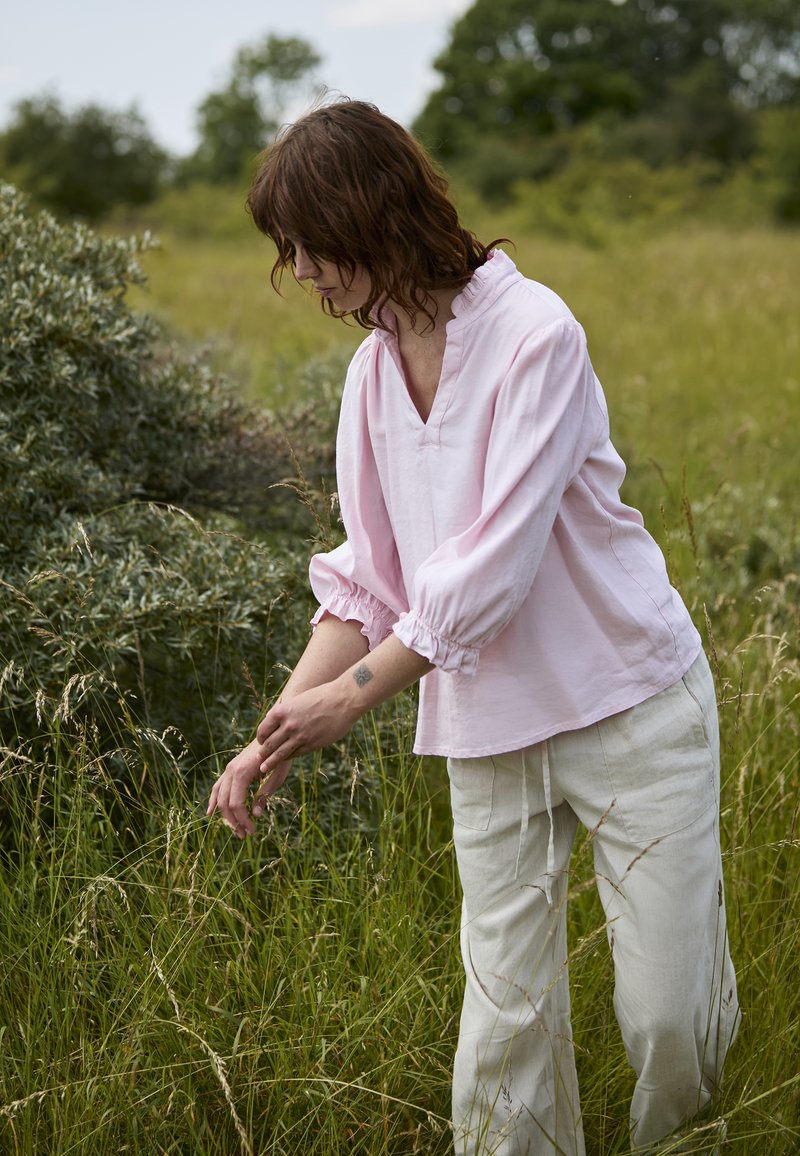 Femme aux cheveux bruns portant un chemisier rose et un pantalon blanc, touchant doucement de l'herbe haute dans un champ vert à l'extérieur.